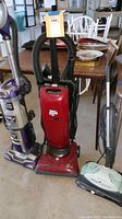 Photo shows three floor cleaning machines: a purple Shark upright vacuum, a red Dirt Devil Jaguar vacuum with hose, and a white Eureka Enviro Steamer with green controls.
