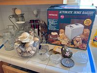 Photo showing Hitachi breadmaker in original box, glass dishes, large glass bowl filled with sea shells, clear glass jars, and kitchen utensils on a kitchen counter.