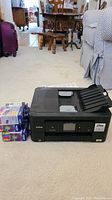 Black Brother Work Smart Series printer with ink cartridges and paper in the background on a carpeted floor.