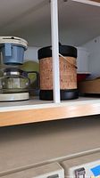 Photo of one beige coffeemaker with blue water reservoir and glass pot, along with a woven ice bucket with black trim and handle on shelf.