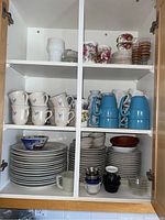 Closed cabinet with two shelves showing stacked white plates, blue insulated mugs, ceramic mugs with blue deer design, floral fine china plates and teacups, and glass bowls.