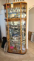 Front angled view of the display cabinet showing four glass shelves inside, wooden frame with rounded edges, glass door, and metal handle. Cabinet contents such as glassware and silverware shown but not included.