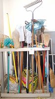 Photo showing multiple garden tools including rakes, shovels, brooms, mop head, and bags of plant food arranged standing in a wooden tool holder against a wall.