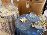 Two glass serving bowls on a floral tablecloth, one clear and one etched with floral design on a silver base, and faux vintage rotary dial telephone with brass and ivory details, glass cup and ladle on a glass platter on a nearby blue cloth draped table.