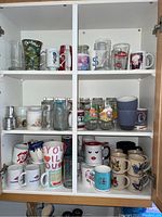 Three shelves inside a cabinet showing an assortment of mugs and glassware including character mugs, souvenir mugs, and clear glasses.