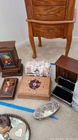 Photo showing seven small jewelry and trinket boxes of varied materials and styles arranged on carpet in front of a wooden cabinet