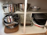 Overview photo showing stack of stainless steel saucepans with black handles on left, large stainless steel pots and mixing bowls on top right, and glass Pyrex rectangular baking dishes in middle bottom shelf along with cooling racks and a brown cutting board.