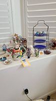 Lot of whimsical ceramics, glass swan, angels, blue hostess tray, and other decorative items on a white surface near window shutters.