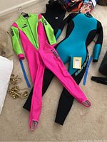Two full wetsuits laid out flat, pink/green SSA wetsuit and teal/black Body Glove wetsuit, with one black vest partially visible