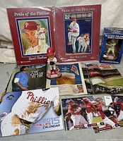 Photo showing Phillies memorabilia assortment including two 'Pride of the Phillies' books featuring Raul Ibañez and Roy Halladay, a pitcher bobblehead, Phillies 2013 calendar, baseball cards, and various printed materials including magazines and congratulatory pamphlets.