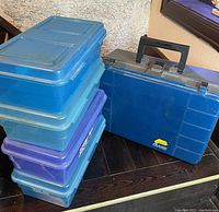 Stack of four plastic storage boxes in blue and purple with a blue Plano tackle box beside them on a dark wood surface.