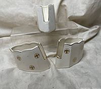Two white porcelain utensil holders with scalloped edges, gold trim, and gold fleur de lis design, placed on cloth background.
