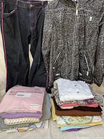 Overview photo showing dark denim pants with pink stitching, black and white patterned zip-up jacket, and assorted folded clothes including a new pink shirt with tag in plastic wrapping.