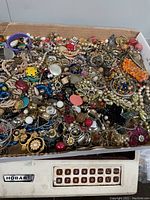 Box filled with a variety of vintage to modern broken and unmatched jewelry pieces, beads, and craft findings showing different colors and styles spread out in a cardboard box.