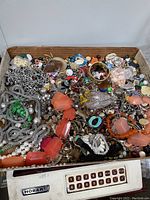 Box filled with assorted broken and unmatched jewelry and craft items, showing a variety of metal chains, beads, rings, and pendants.