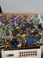 Photo showing a wide mix of broken vintage to modern jewelry pieces, including faux pearl necklaces, colorful beads, metal chains, and ribbons piled in a box on a scale.