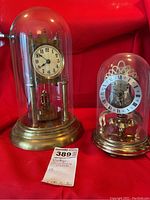 Two anniversary dome clocks: one taller with solid brass base and Arabic numeral dial, one smaller with brass finish base and Roman numeral dial, both under glass domes on a red background.