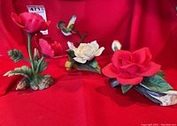 Three ceramic flower figurines: two red poppy-like flowers and one white flower with a hummingbird on a branch, displayed against a red backdrop.