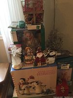 Multiple Christmas decoration items arranged on small table near window with natural light, including figurines, boxed decorations, decorative wire tree, and light boxes.