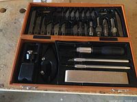 Full overview of the Exacto cutting tool set inside the open brown storage case, showing blades, handles, sharpening stone, and clamps.