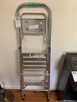Full view of folded silver metal step ladder with visible wear and green tape on the top rail.