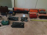 Overview of vintage Lionel Scout train set components including locomotive, tender, boxcars in original orange boxes, red caboose, curved and switch track pieces, and wiring.