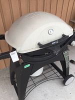 Front and side views of Weber Q gas grill with cream white hood on black plastic stand. Includes temperature gauge and control knobs, wheels for mobility.