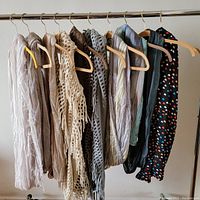 Ten infinity scarves displayed hanging on hangers showing various materials, textures, and styles from neutral and pastel colors to dark black prints.