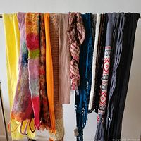 Photo showing 12 scarves hanging side by side including yellow knit, multicolor felt, orange knit, beige knit, light brown knit with dots, ruffled multicolor, blue knit, black knit, patterned black and white knit, gray cable knit, white with red and black pattern knit, and black ribbed knit.