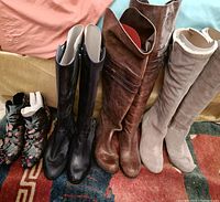 Four pairs of women's boots in various colors and heights placed on a rug and fabric backdrop, showing condition and details including zippers, stitches, embroidery, and lining.
