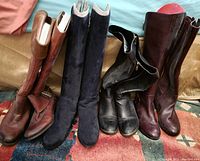 Four pairs of women's tall boots arranged side by side on a carpeted floor, showing various colors and styles including brown leather with buckle, black suede, black leather with zipper, and dark reddish-brown leather.