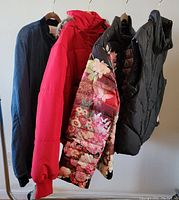 Four women's jackets and vests hanging on a rack, including a navy jacket, red puffer jacket with fur hood, floral jacket, and black vest