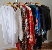 Photo showing seven women's wraps and ponchos hanging on wooden hangers, with various colors and patterns including white, geometric beige, red floral, blue floral, and black semi-sheer