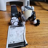 Photo of Chip robot dog lying on wooden floor with charging dock, ball, two watches, and instruction manual arranged around it.
