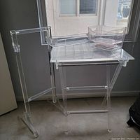 Photo of a group lot showing two acrylic folding trays with foldable legs, an acrylic valet stand, and two rectangular acrylic organizing bins with curved wave designs on the short sides, all made of clear transparent acrylic, placed indoors near a window.
