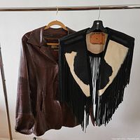 Front view of brown Lucien Piccard leather jacket and custom cowhide fringe vest hanging on rack