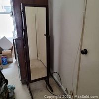 Full view of standing floor wooden jewelry cabinet with full-length mirror on door, showing cabinet legs and room environment.