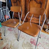 Photo showing two light brown vinyl folding chairs with chrome tubular frames from the side angle, displaying decent condition.