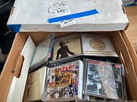Cardboard box with assorted empty CD jewel cases, some containing front cover paper inserts of music albums but no discs inside.
