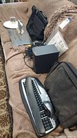 Photo showing Logitech keyboard, laptop bags, Dell speaker subwoofer, power adapter and some cables on a couch.