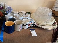 Photo showing assortment of mugs in various colors and designs, stacked plates including turkey decorated serving plate, and plastic cake plate with clear cover on top, placed on a wooden surface next to artificial flowers.