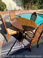 Photo of oval wooden dining table with 5 dining chairs around it, partly shaded outdoors near pool