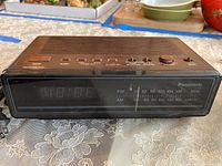 Front view of a Panasonic clock radio, digital display off, speaker grille on top, various control buttons and dials visible.