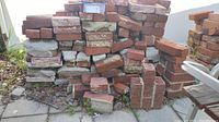 Stack of used red pavestone bricks, showing various bricks with worn edges, some with mortar remnants, stacked on a paved surface with outdoor background.