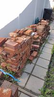 View of large stacks of red bricks arranged in rows along a paved path next to a fence, showing the quantity and condition of the bricks.