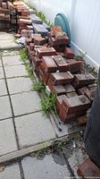 Two piles of red bricks stacked outdoors, some bricks broken or chipped. Piled on ground near white fence and slabs.