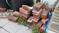 Photo shows stacked red pavestone bricks of various conditions, some broken and weathered, stored outdoors on concrete slabs next to a fence and garden soil