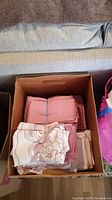 Open cardboard box containing neatly folded rose pink cloth napkins and beige/ivory napkins, some with decorative lace edges.
