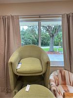 Green fabric chair shown upright with a cushioned seat in front of a window with beige curtains.