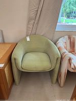 Green fabric chair by Directional shown from front angle, placed next to a wooden table and another chair, against beige walls.